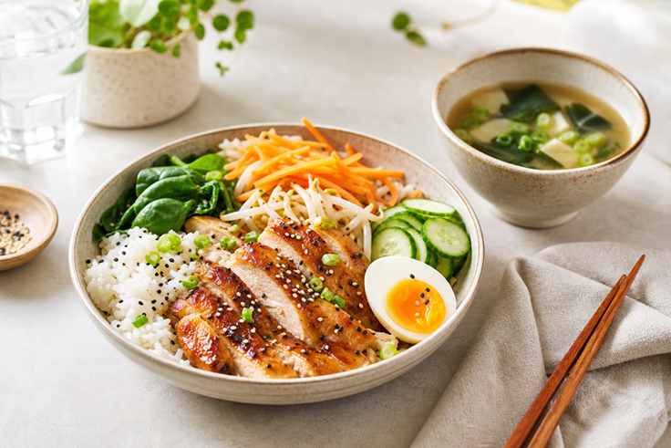 A plated lunch bowl with rice, grilled chicken, vegetables, egg, and soup